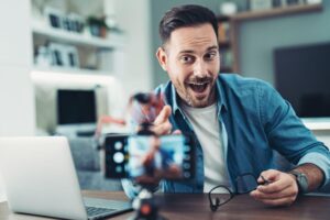 A cheerful content creator recording a video at home with a smartphone camera and laptop, representing the future of digital marketing, online content creation, and influencer branding